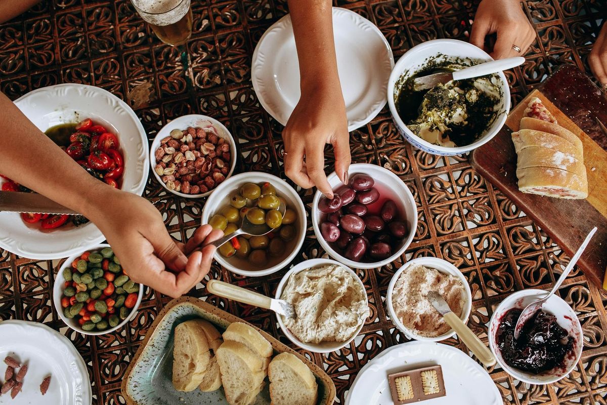 Overhead view of a rustic Mediterranean meal with diverse dishes and hands reaching across the table.