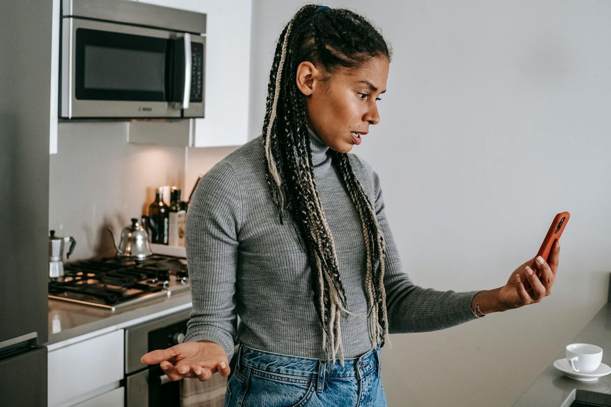 Serious young black lady with Afro braids in casual clothes gesticulating while having unpleasant conversation via video chat on smartphone in modern kitchen.