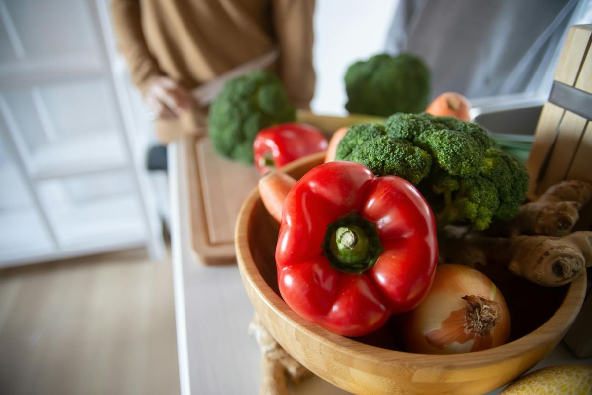 A close-up of fresh vegetables including red peppers, broccoli, and ginger in a wooden bowl.