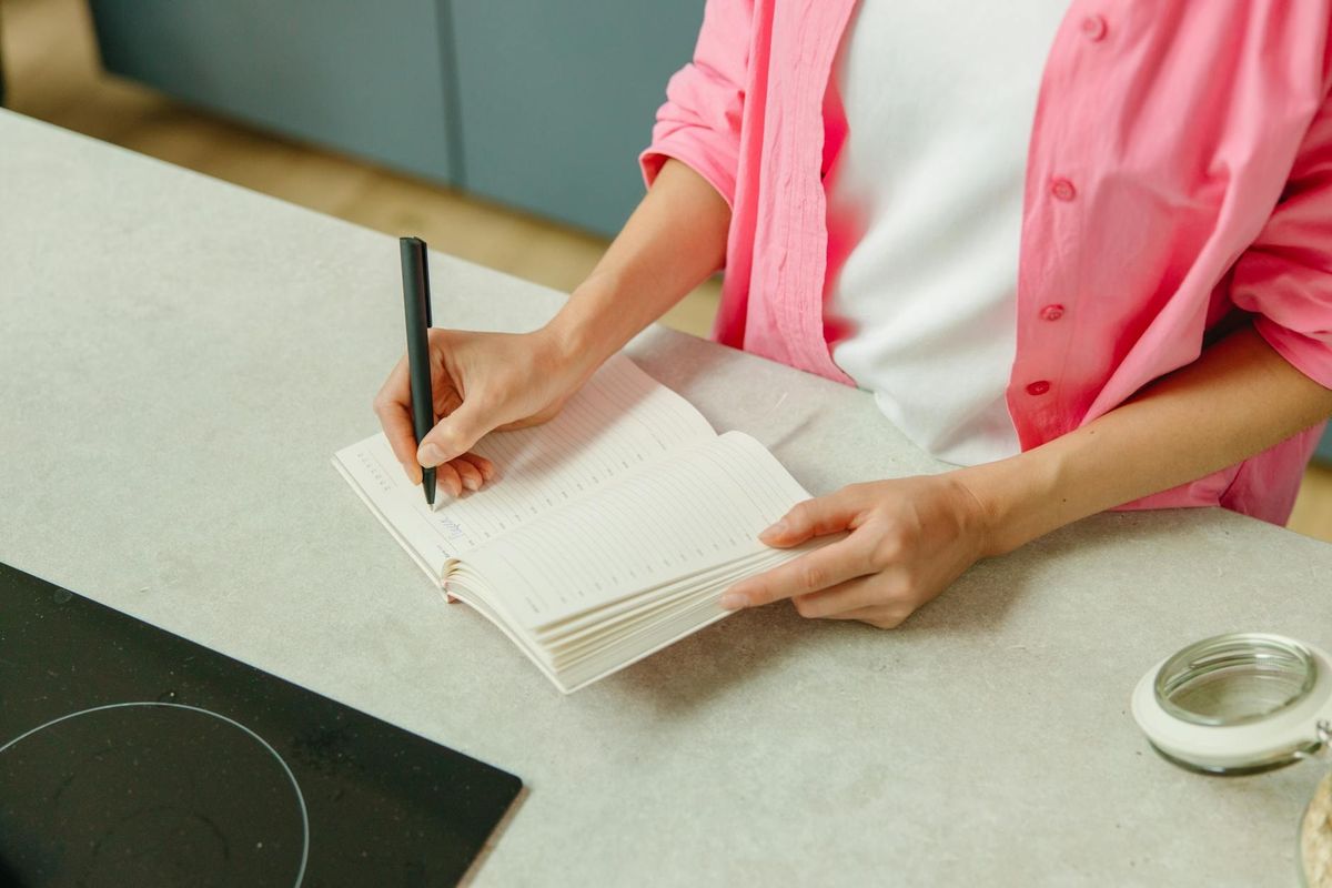 A woman in a pink shirt writing in a notebook on a kitchen counter with a pen.